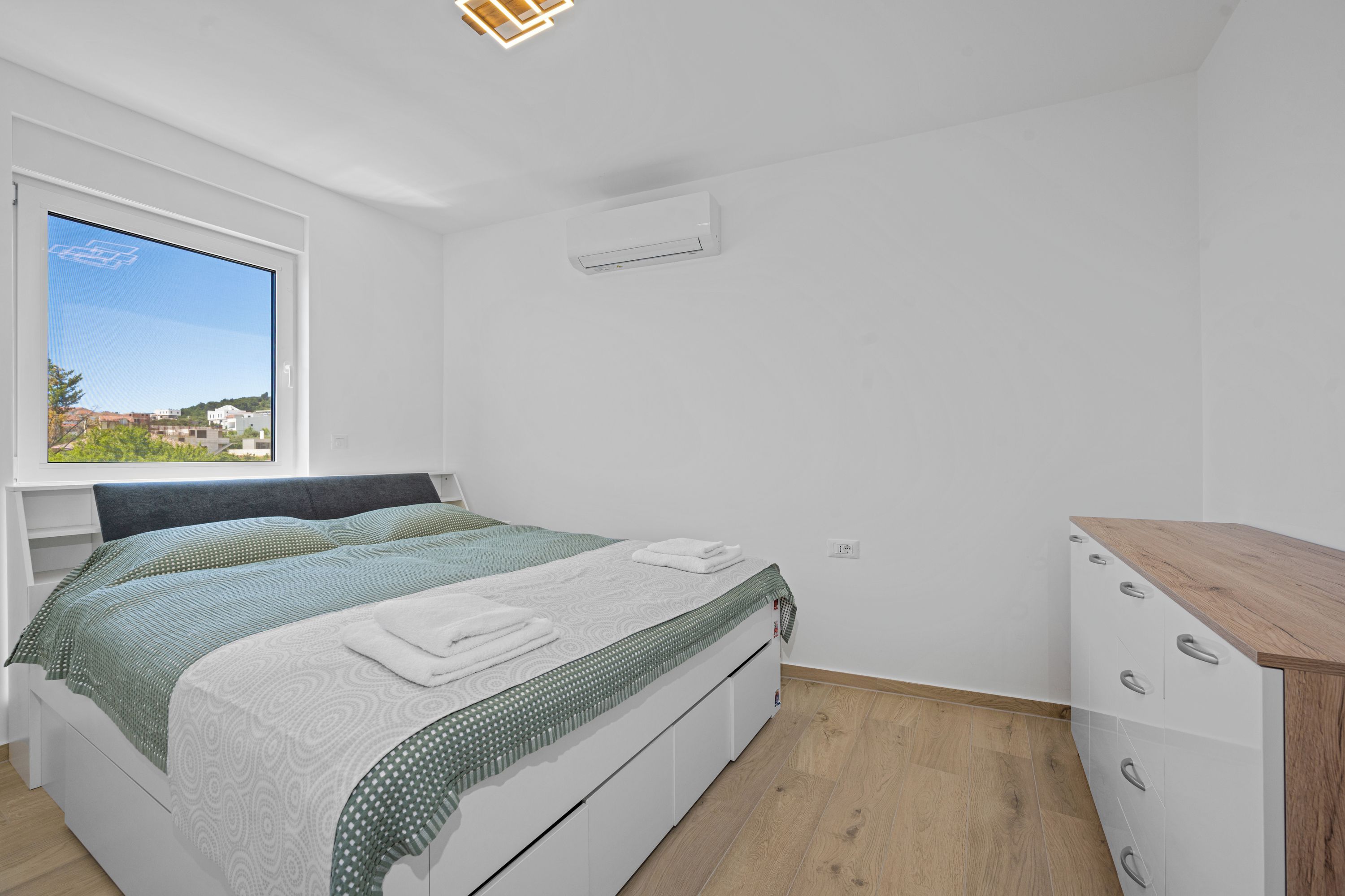 A bedroom with a bed, a window with a view, a dresser, and an air conditioning unit.