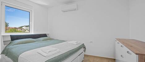 A bedroom with a bed, a window with a view, a dresser, and an air conditioning unit.