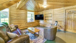 Interior de una cabaña de madera con un sofá de cuero, una mesa de centro, un otoman redondo, un televisor y una puerta.