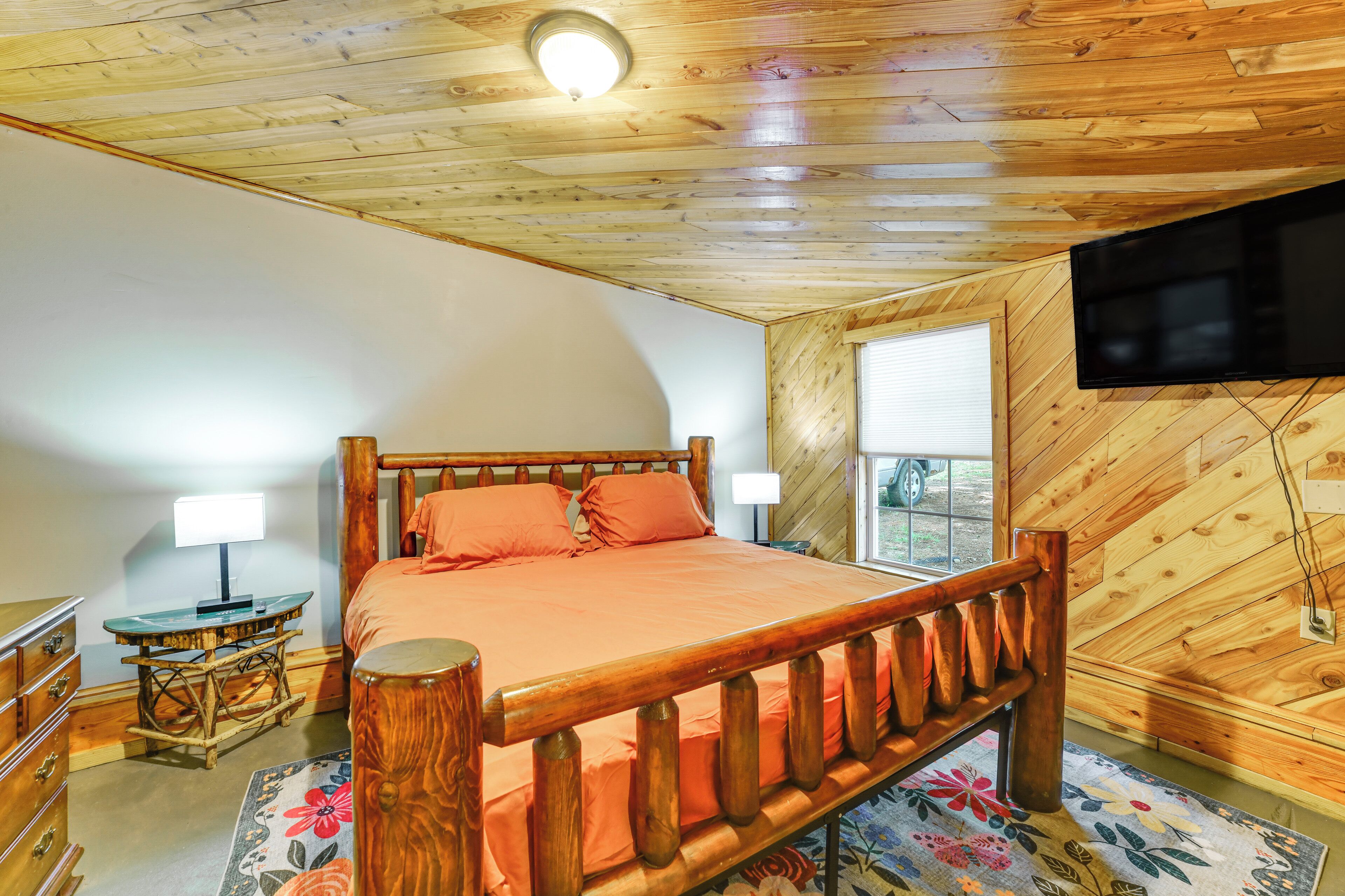 Une chambre avec un plafond en bois, un lit avec une literie orange, une table de chevet en bois, un bureau avec une lampe et un téléviseur fixé au mur.