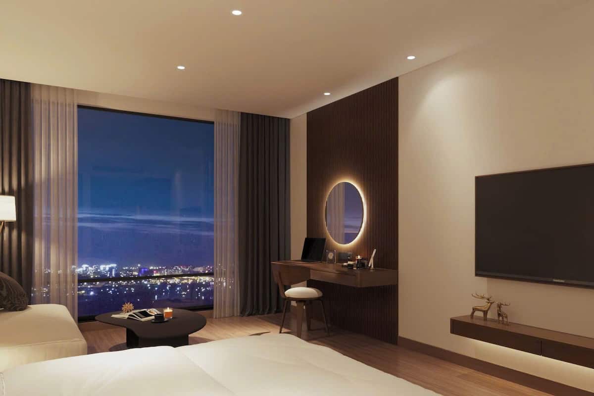 A modern hotel room with a large bed, a desk with a laptop, a TV, and a view of a cityscape at night.