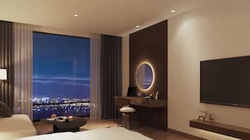A modern hotel room with a large bed, a desk with a laptop, a TV, and a view of a cityscape at night.