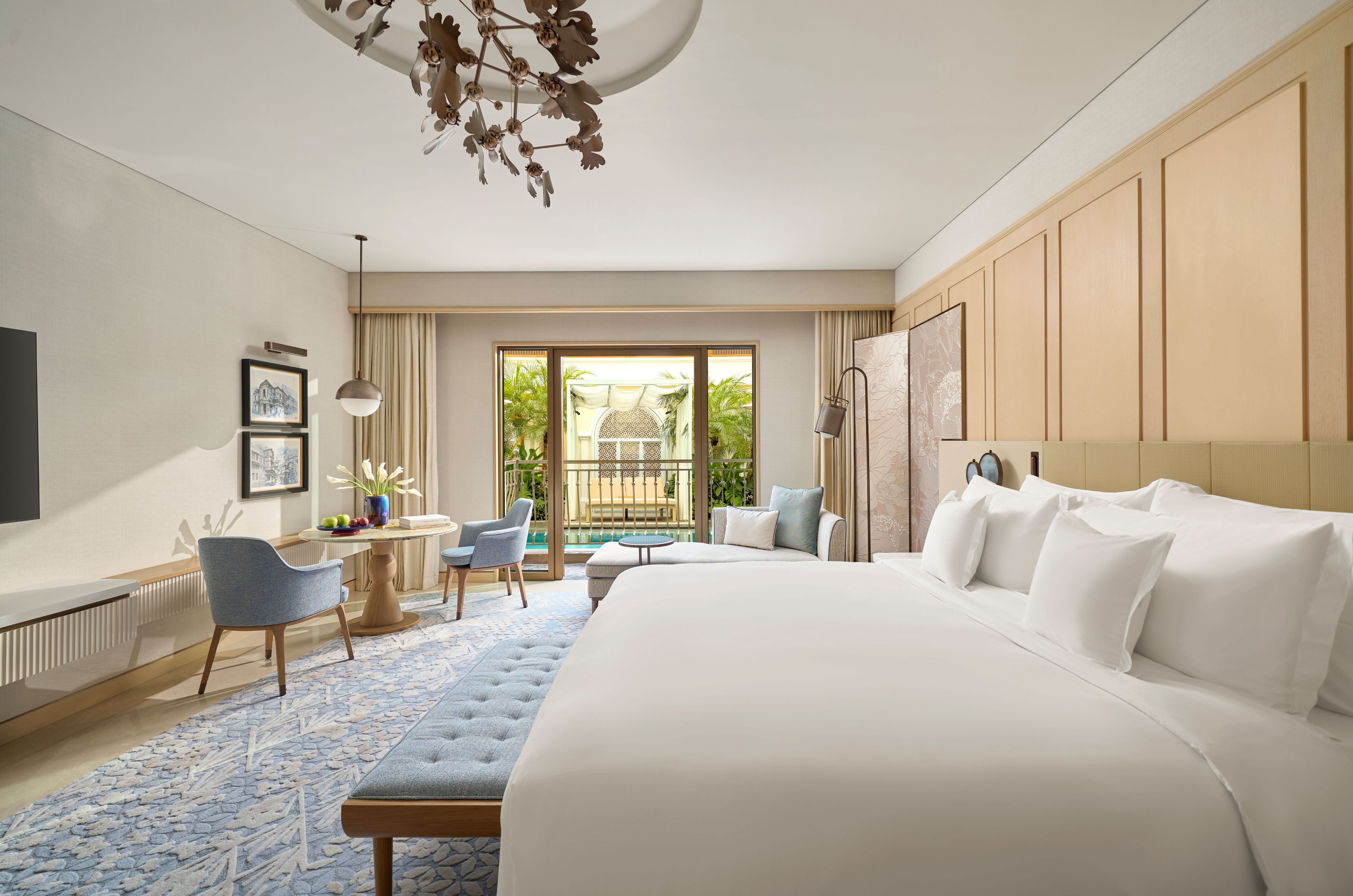A hotel room with a large bed, a desk with a chair, a small table with a chair, a large window with a view of greenery, and a ceiling light fixture.