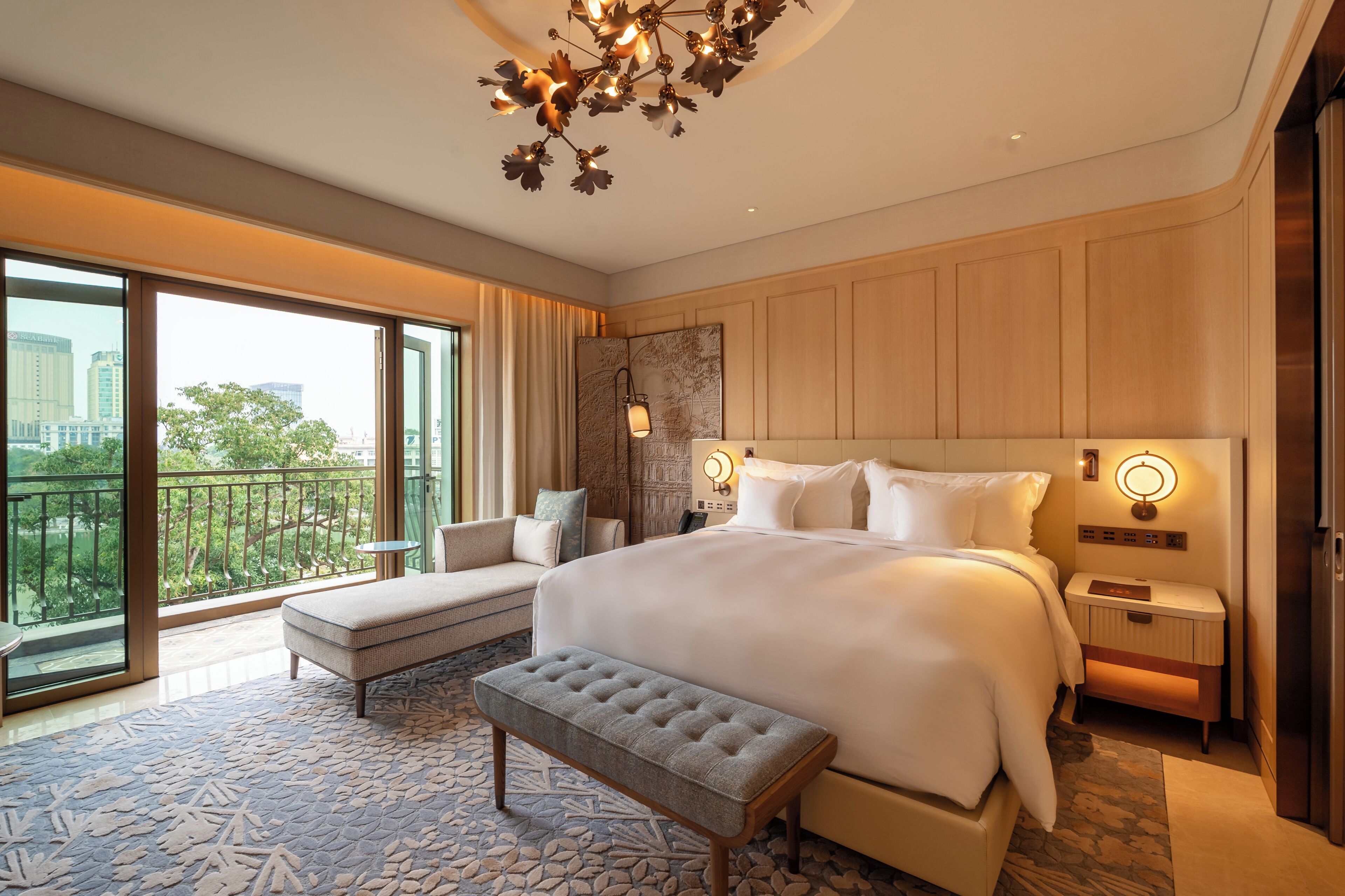 A hotel room with a large bed, a sitting area with a bench, a view of the city through the balcony, and a modern chandelier.