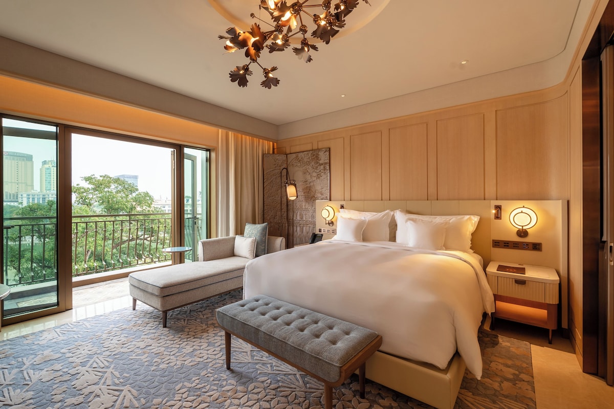 A hotel room with a large bed, a sitting area with a bench, a view of the city through the balcony, and a modern chandelier.