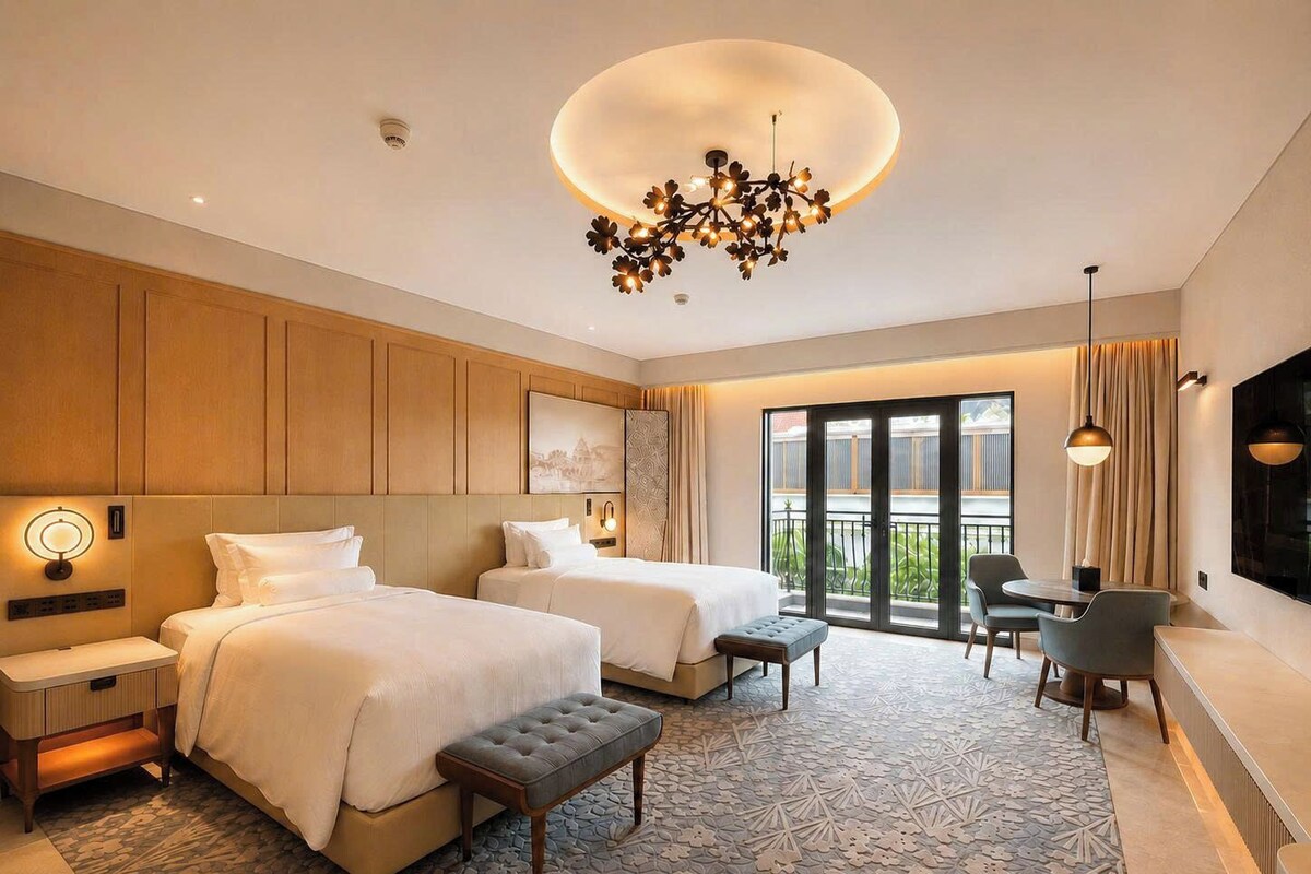 A hotel room with two beds, a sitting area with a table and chairs, a balcony with a view, and a modern chandelier.