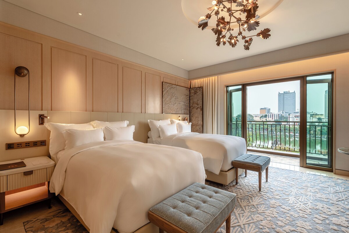 A hotel room with two beds, a nightstand, a bench, a large window with a city view, and a decorative ceiling light.