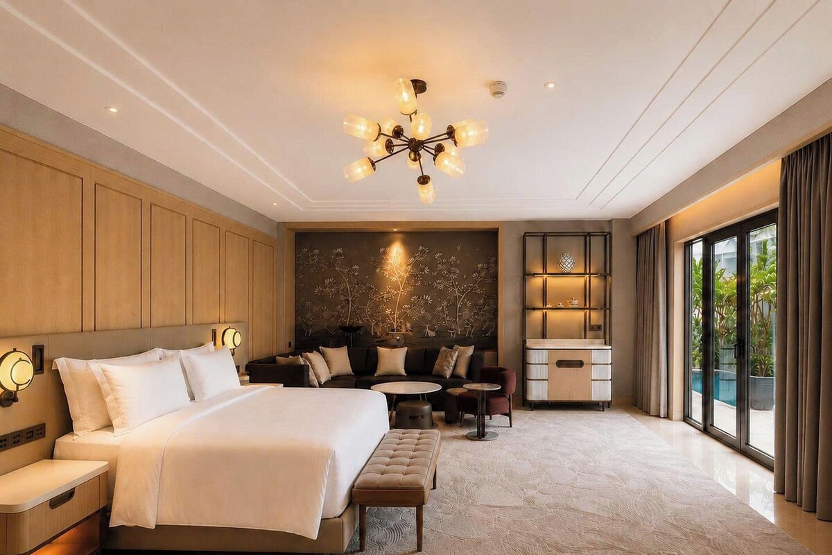 A modern hotel room with a large bed, seating area, and a view of greenery outside.