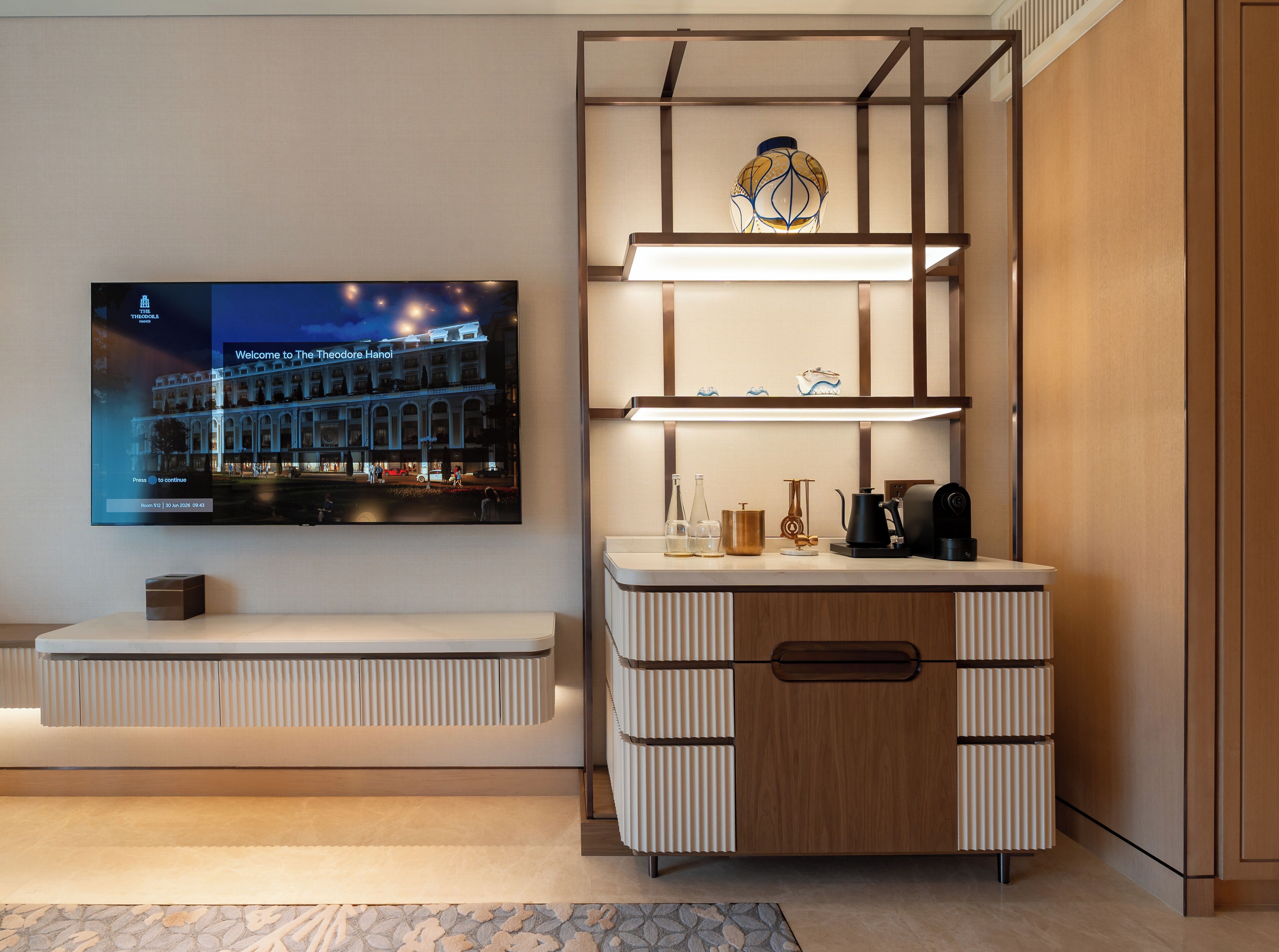 A modern hotel room with a flat-screen TV displaying a welcome message, a built-in shelf with decorative items, a wooden cabinet with a drawer, and a patterned rug.
