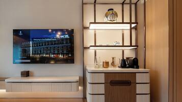 A modern hotel room with a flat-screen TV displaying a welcome message, a built-in shelf with decorative items, a wooden cabinet with a drawer, and a patterned rug.