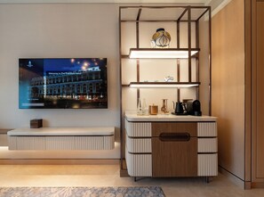 Una camera d'albergo moderna con una TV a schermo piatto che mostra un messaggio di benvenuto, una mensola a incasso con oggetti decorativi, un mobiletto in legno con un cassetto e un tappeto a motivi.