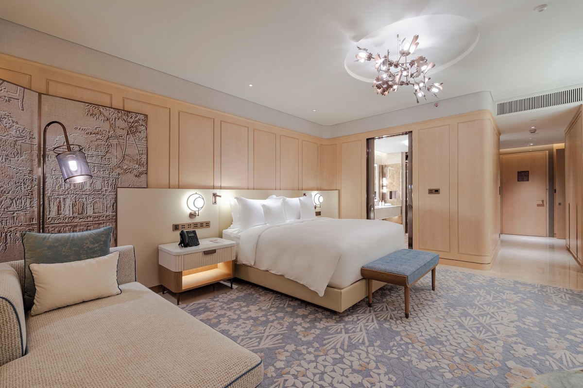 A modern bedroom with a large bed, bedside lamps, a nightstand, a bench, and a decorative wall piece.