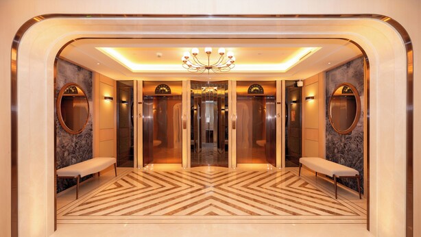 A modern elevator lobby with a geometric patterned floor, two benches, a chandelier, and mirrored walls. - The Theodore Hanoi (Hanoi)
