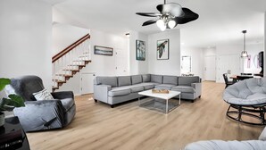Een moderne woonkamer met een grijze hoekbank, een ronde fauteuil, een salontafel, een trap en een plafondventilator.