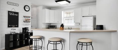 A modern kitchen with white cabinetry, a central island with stools, a coffee bar sign, and a welcome sign.