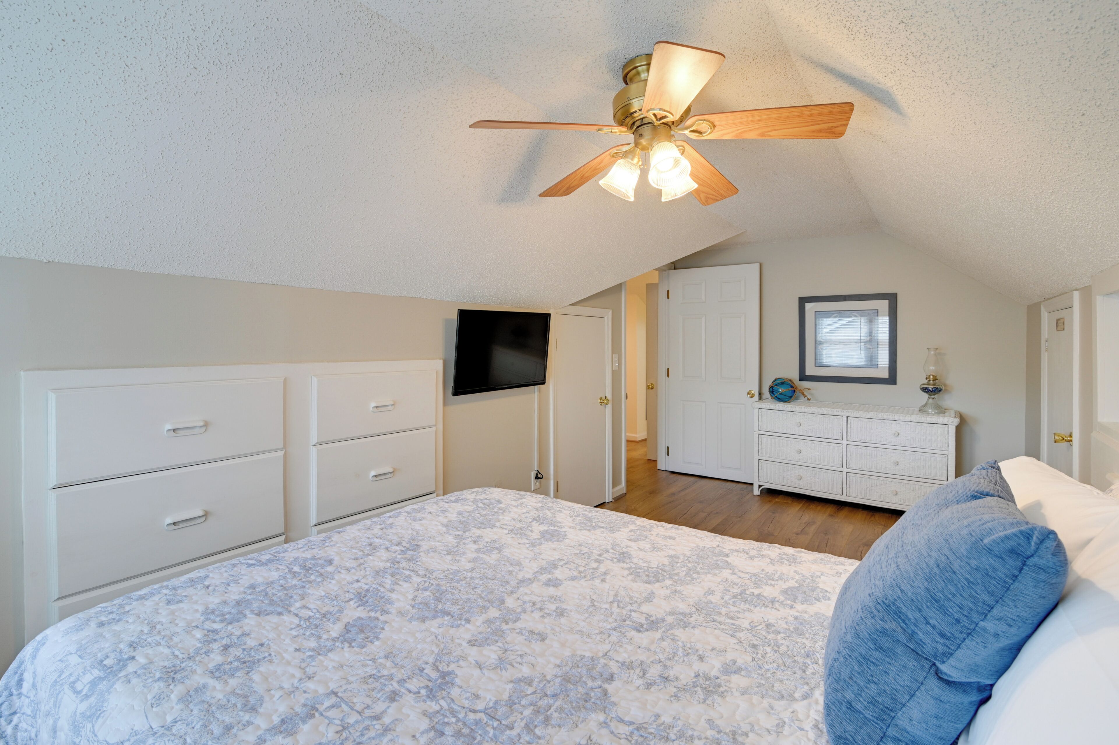 A bedroom with a bed, a ceiling fan, a dresser, a television, and a door leading to another room.