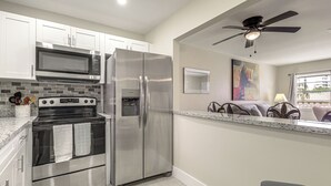 Una cocina con electrodomésticos de acero inoxidable, gabinetes blancos, salpicadero de azulejos y ventilador de techo.