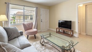 Una sala de estar con sofá, sillón, mesa de centro de vidrio, televisor y puerta que da a un balcón.
