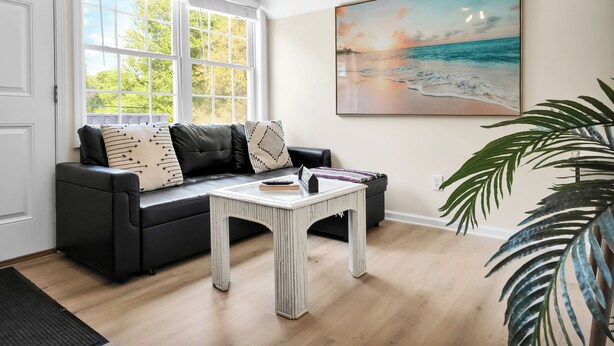 Uma sala de estar com um sofá preto, uma mesa de centro branca, uma janela grande e um quadro com uma paisagem praiana.