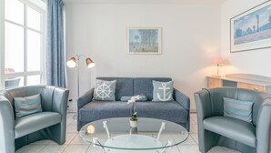 Ein modernes Wohnzimmer mit einem grauen Sofa, zwei Sesseln, einem Glastisch und einer Lampe.