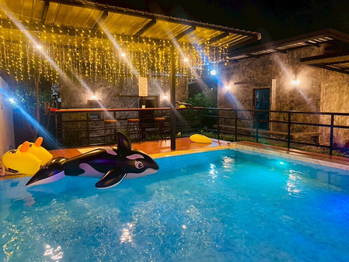 Uma piscina com brinquedos flutuantes de orca, área de bar e luzes de corda.
