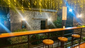 Een buitengebied met een bar, een stenen gebouw, hangende lichten en uitzicht op groen.