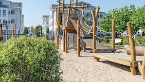Eine hölzerne Spielgerüst mit einer Seilbrücke, umgeben von Sand und Grünflächen, mit Gebäuden im Hintergrund.