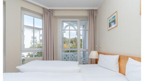 Hotellihuone, jossa on kaksi sänkyä, ikkuna, jossa on verhot, lamppu ja seinällä oleva kehystetty kuva.