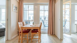 A dining area with a wooden table and chairs, large windows with curtains, and a view of buildings outside. - Ferienwohnung im 2.OG mit Balkon in Sellin (Sellin)