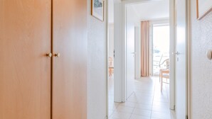 Un couloir avec une porte en bois, des murs blancs et une vue sur une pièce où l’on aperçoit une chaise et des rideaux.