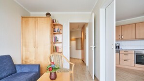 Un salon comprenant un canapé, une table en bois avec un vase, une bibliothèque et un coin cuisine avec des placards et des appareils électroménagers.