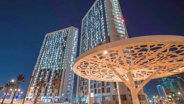 Deux immeubles de grande hauteur dont les fenêtres sont illuminées la nuit, une structure métallique décorative au premier plan et des palmiers bordant la rue.