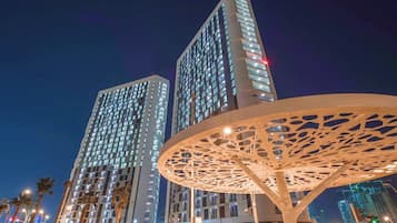 Deux immeubles de grande hauteur dont les fenêtres sont illuminées la nuit, une structure métallique décorative au premier plan et des palmiers bordant la rue.