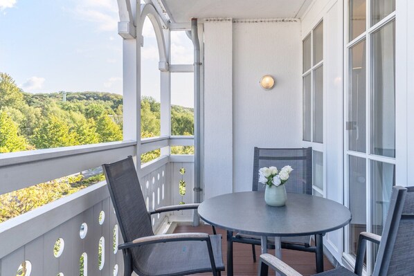 En balkon med et rundt bord og stole, en vase med blomster, og udsigt til en skovklædt bakke.