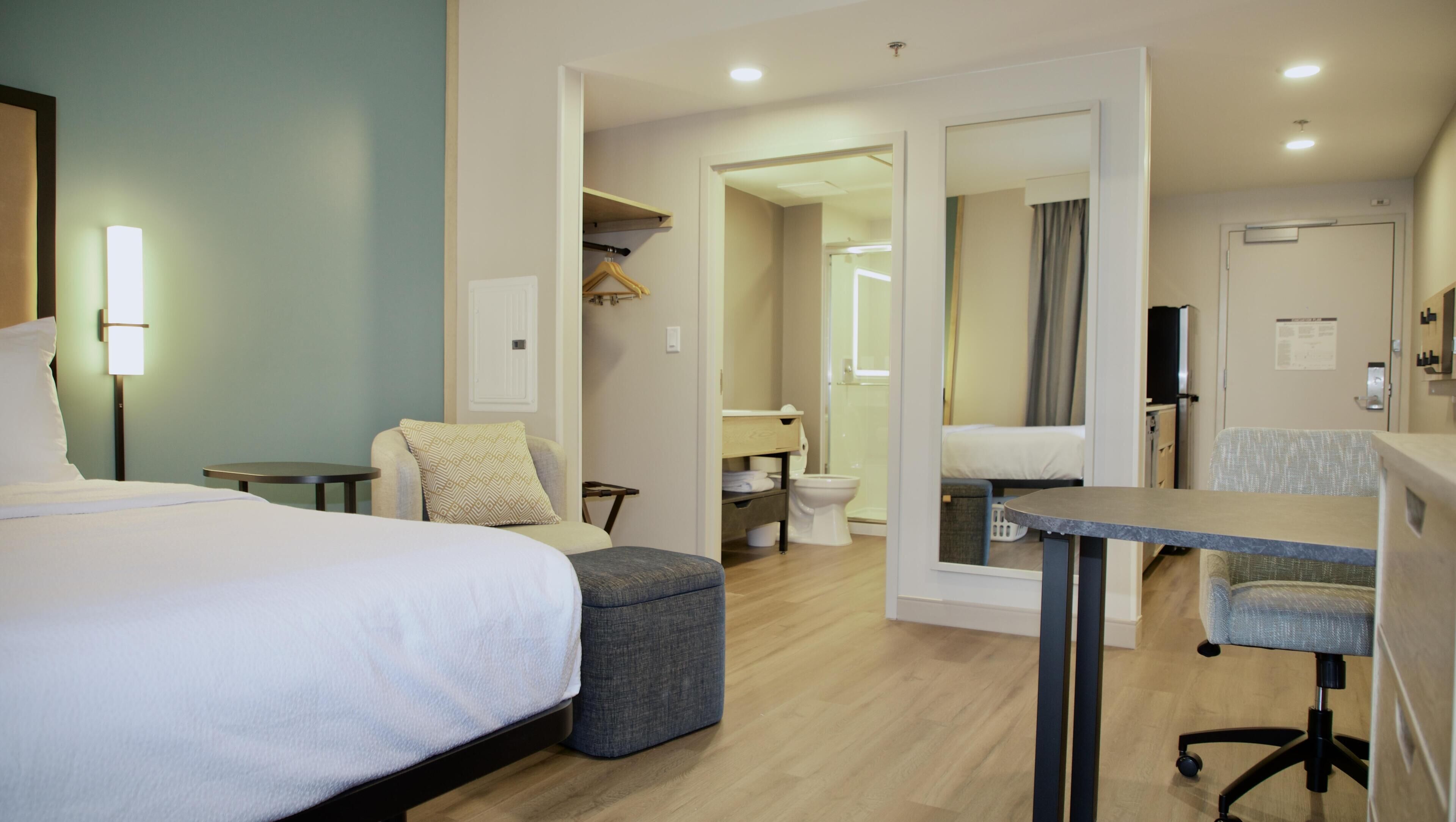 Une chambre d’hôtel avec un lit, un bureau, une chaise, une petite table, un miroir et une salle de bain visible à travers une porte ouverte.