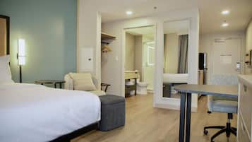 Une chambre d’hôtel avec un lit, un bureau, une chaise, une petite table, un miroir et une salle de bain visible à travers une porte ouverte.