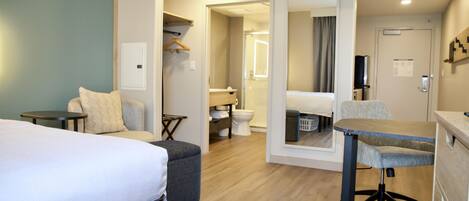 Habitación de hotel con cama, escritorio, silla, baño con inodoro y ducha, y espejo.