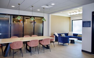 A modern meeting room with a long table, several chairs, a blue sofa, and a wall-mounted screen.