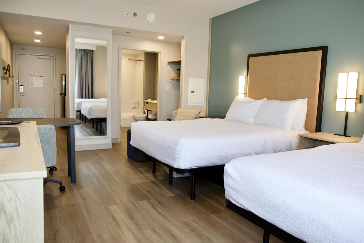 Habitación de hotel con dos camas, un escritorio, una silla, baño y zona de ducha.