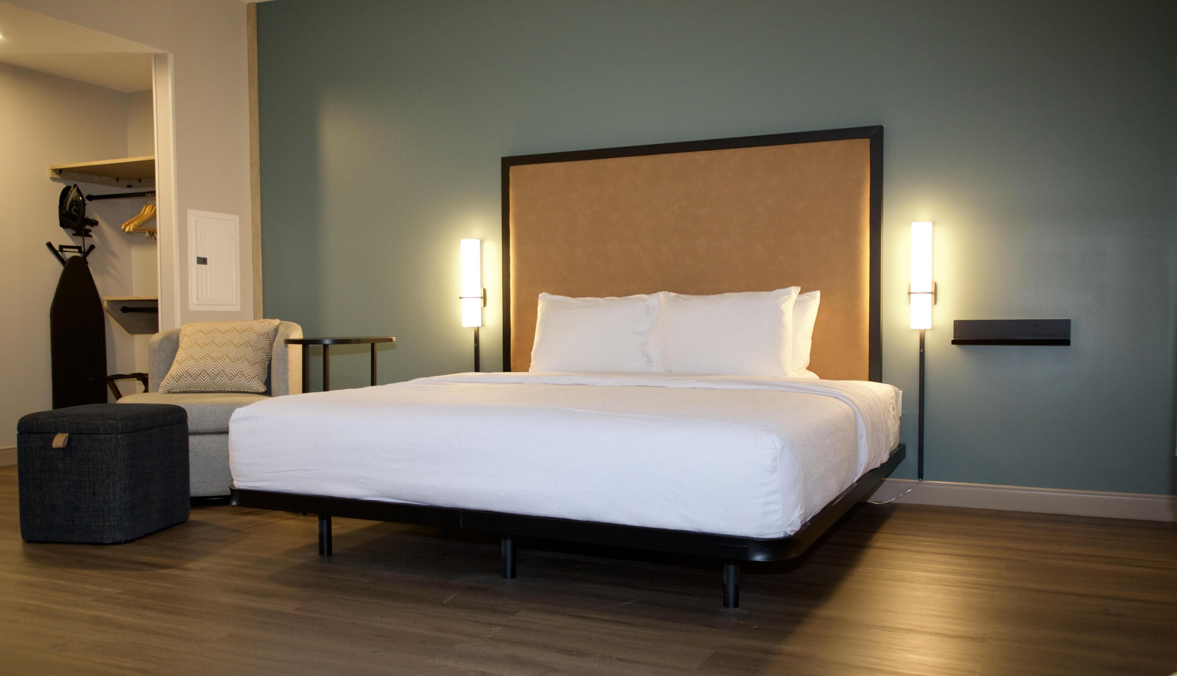 Habitación de hotel con una cama grande, dos lámparas de noche, una mesita, una silla y un perchero.