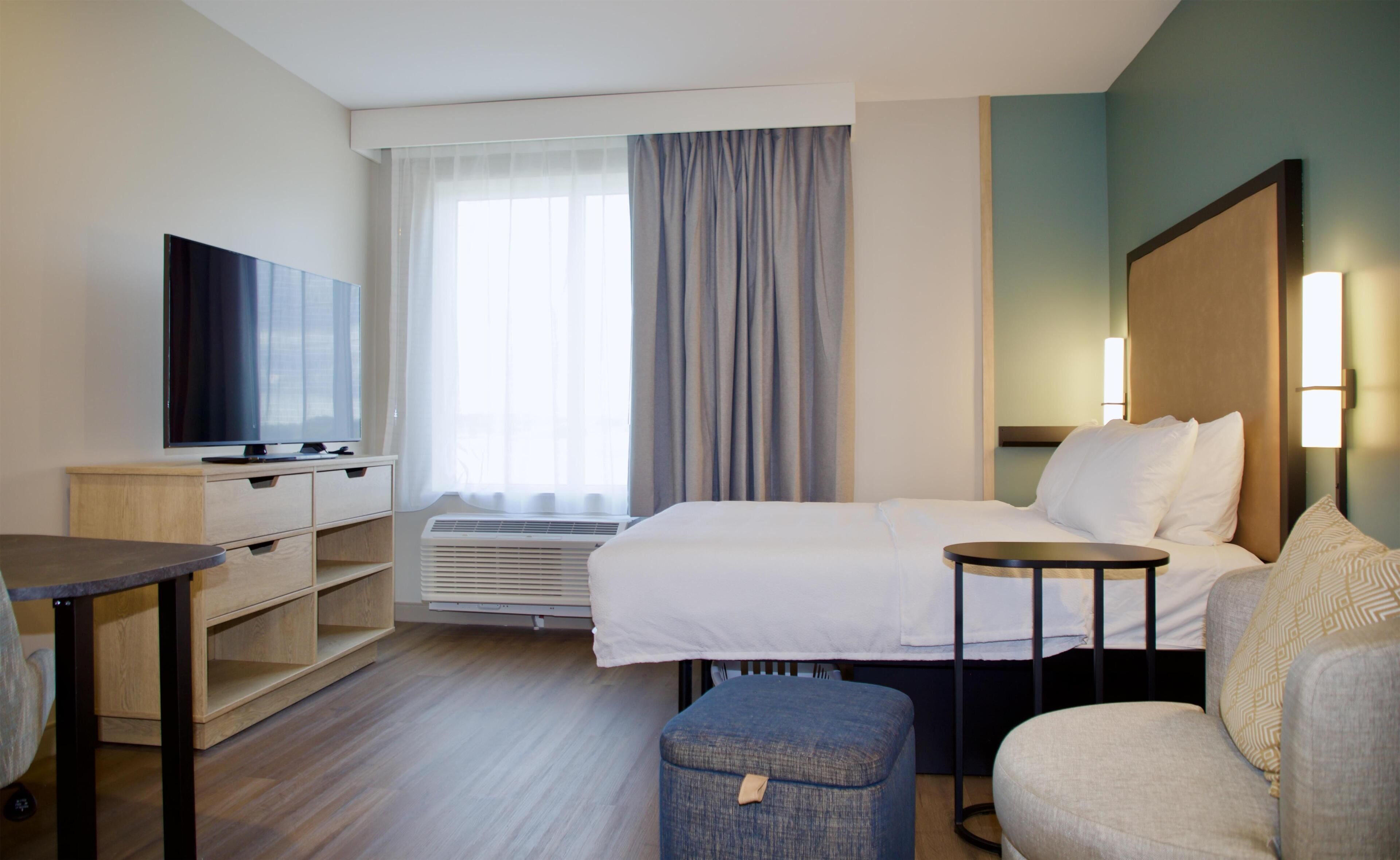 Et hotelværelse med seng, skrivebord, fjernsyn, et vindue med gardiner og et sengebord.