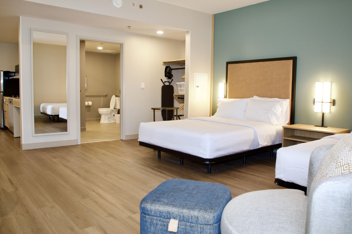 Habitación de hotel con una cama grande, una cama más pequeña, un escritorio, una silla y un baño visible a través de una puerta abierta.