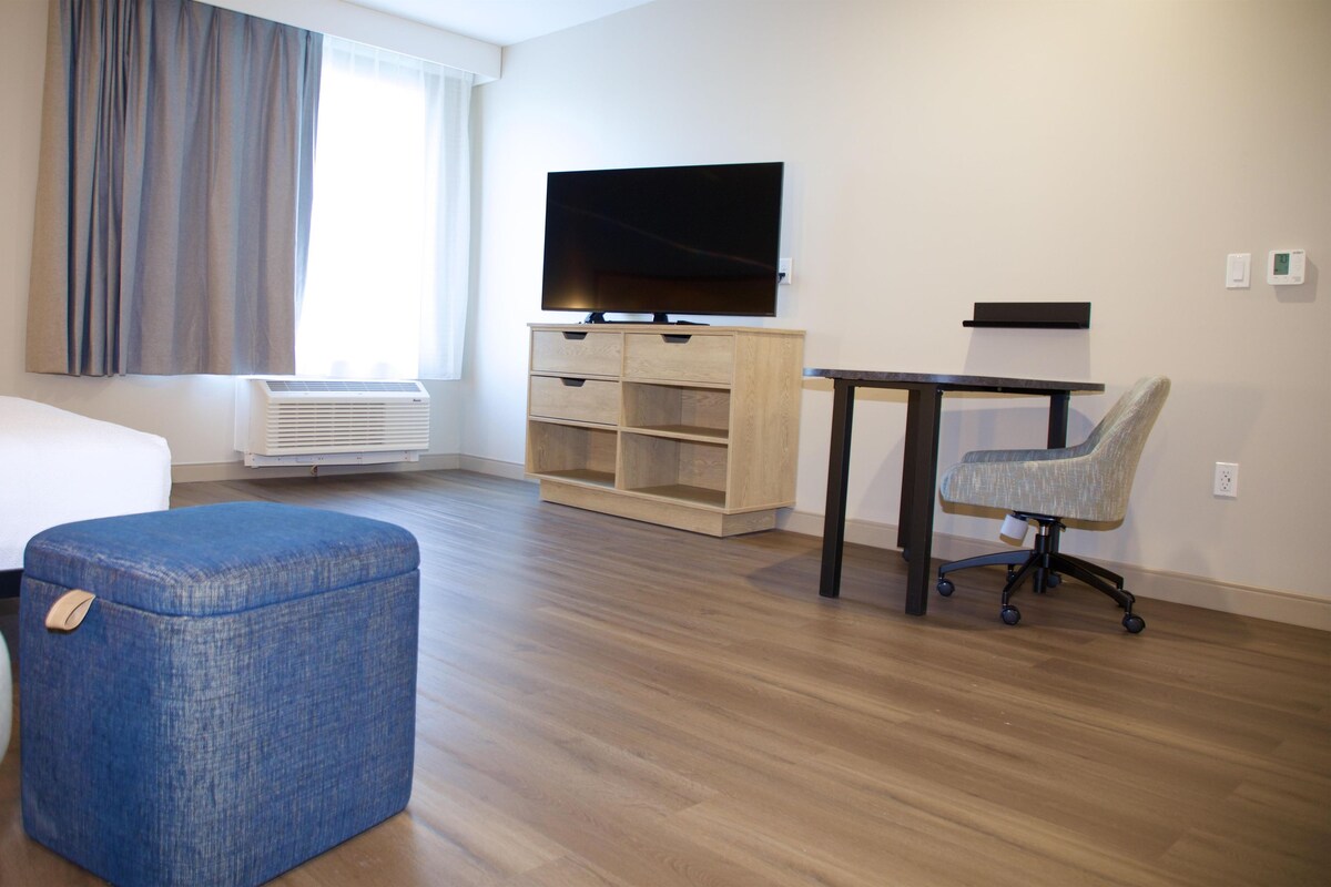 Habitación de hotel con cama, escritorio, silla, televisor y ventana con cortinas.