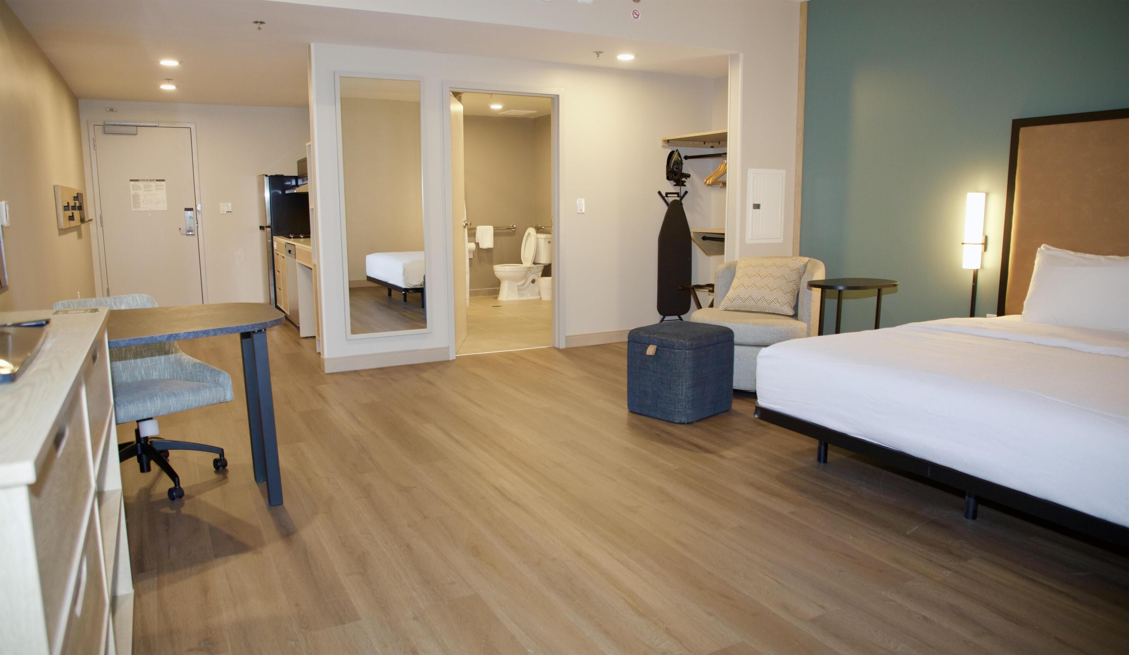 A hotel room with a bed, a desk, a chair, a suitcase, and a bathroom visible through an open door.
