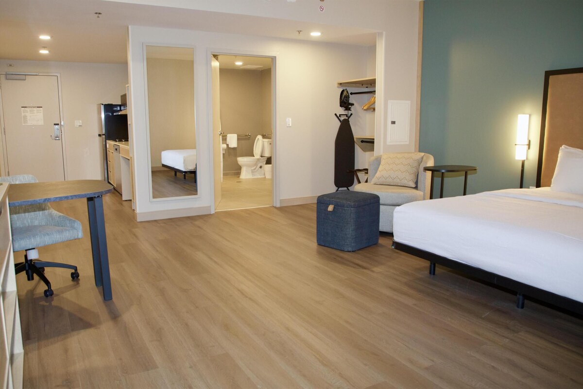 A hotel room with a bed, a desk, a chair, a suitcase, and a bathroom visible through an open door.