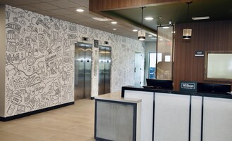 Zona de recepción de hotel con una pared decorada con dibujos, dos ascensores y un letrero que indica la oficina del gerente.