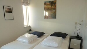Een tweepersoonsbed met witte lakens, een nachtkastje met lamp, een klein tafeltje met een stoel, en een schilderij aan de muur.