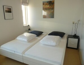 Ein Doppelbett mit weißer Bettwäsche, ein Nachttisch mit Lampe, ein kleiner Tisch mit Stuhl und ein Bild an der Wand.
