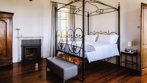 A four-poster bed with a bench, a fireplace, and a nightstand with a lamp.