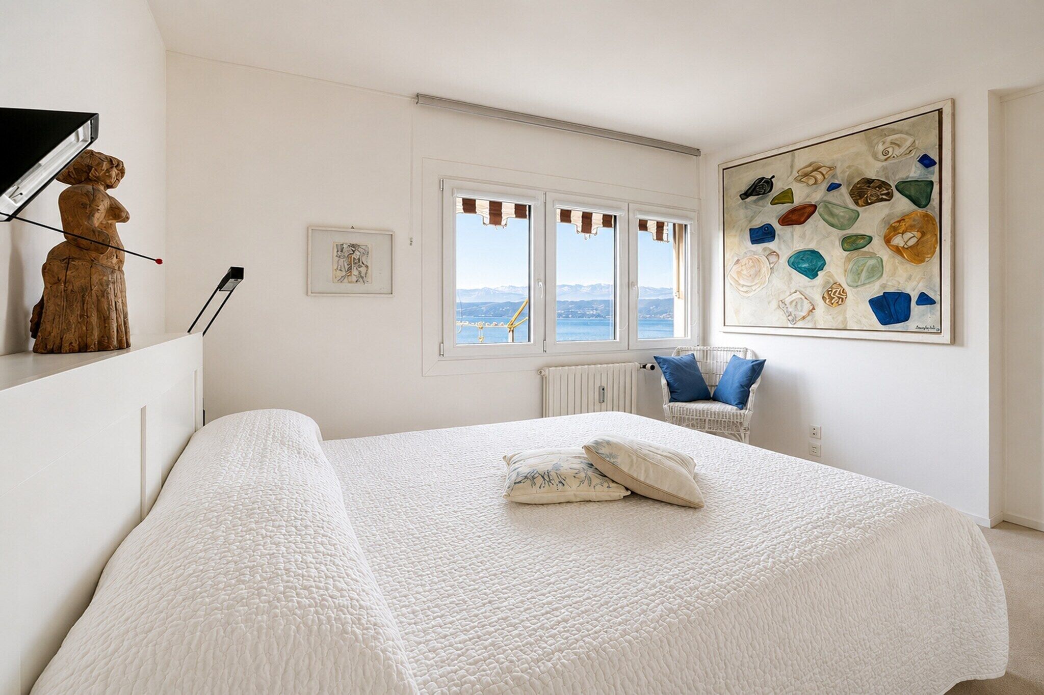 A bedroom with a large bed, a view of the sea through windows, a wooden sculpture, and a painting.